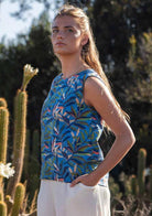 Model posing side ways in front of cactus plants wearing 100 percent cotton blue leafy print sleeveless top with high round neckline and hand in pants pocket