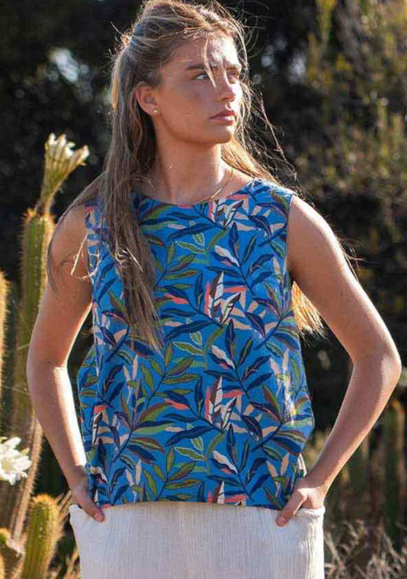 Model standing in front of cactus plants looking sideways wearing 100 percent cotton blue leafy print sleeveless top with high round neckline and hands in pants pockets