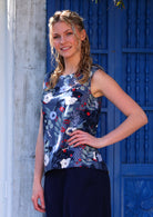 Model standing sideways in front of blue door with hand on hip wearing 100 percent cotton navy blue floral sleeveless top that is slightly higher at the front