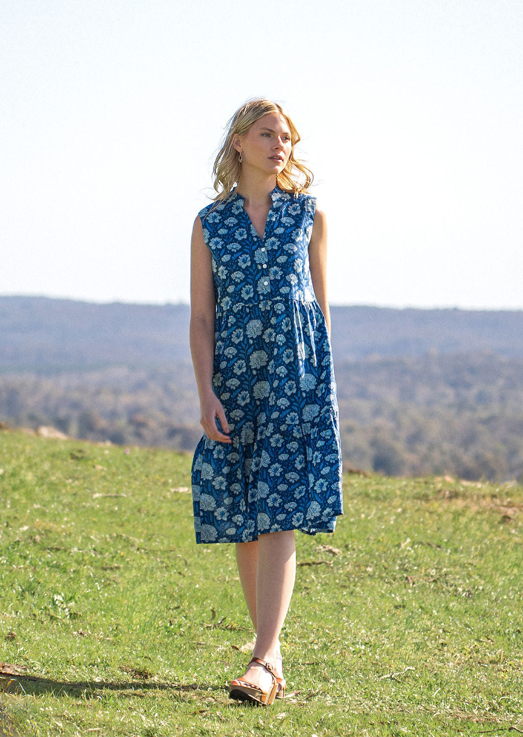 Woman walks across field in cotton sleeveless midi length dress with two tiered skirt in blue floral print