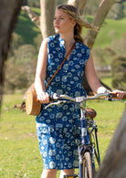 Woman stands with bicycle in sleeveless cotton midi length dress with three buttons in bodice and mandarin collar