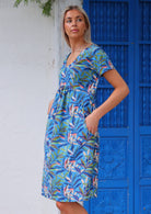 Woman stands sideways in blue based leafy print cotton dress with an empire waistline and her hand in pocket