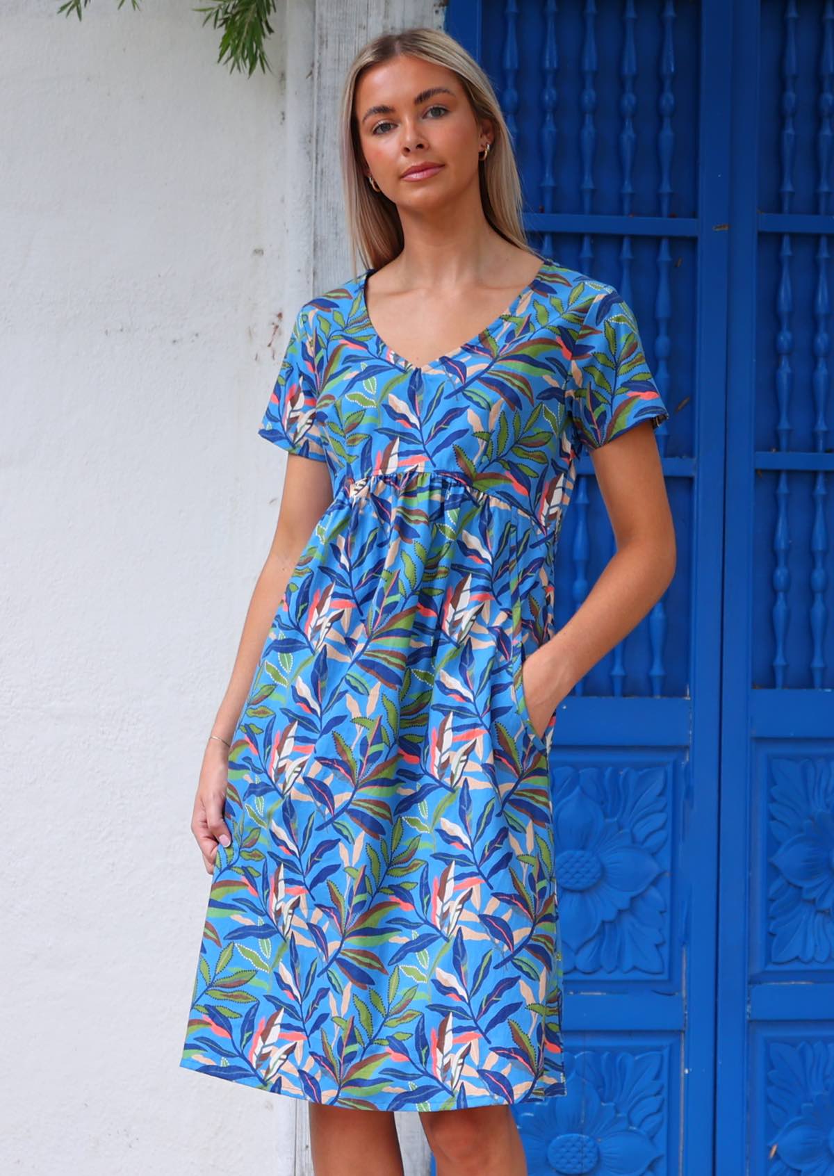 Model poses in cotton over the knee dress in blue based leafy print in multi-colours with her hand in pocket