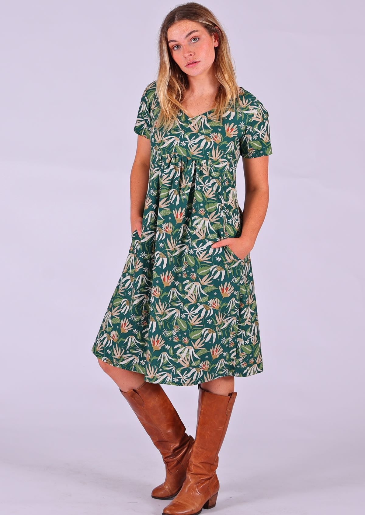 Model standing with hands in pockets modelling the Etta v-neck Dress Flannel Flower 100 percent cotton green sundress with short sleeves paired with tan leather boots