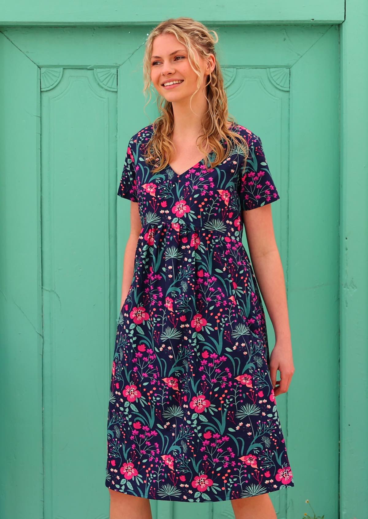 Model wearing 100 percent cotton v-neck sundress with hot pink and bright purple flowers on a deep blue base, with one hand in pocket 