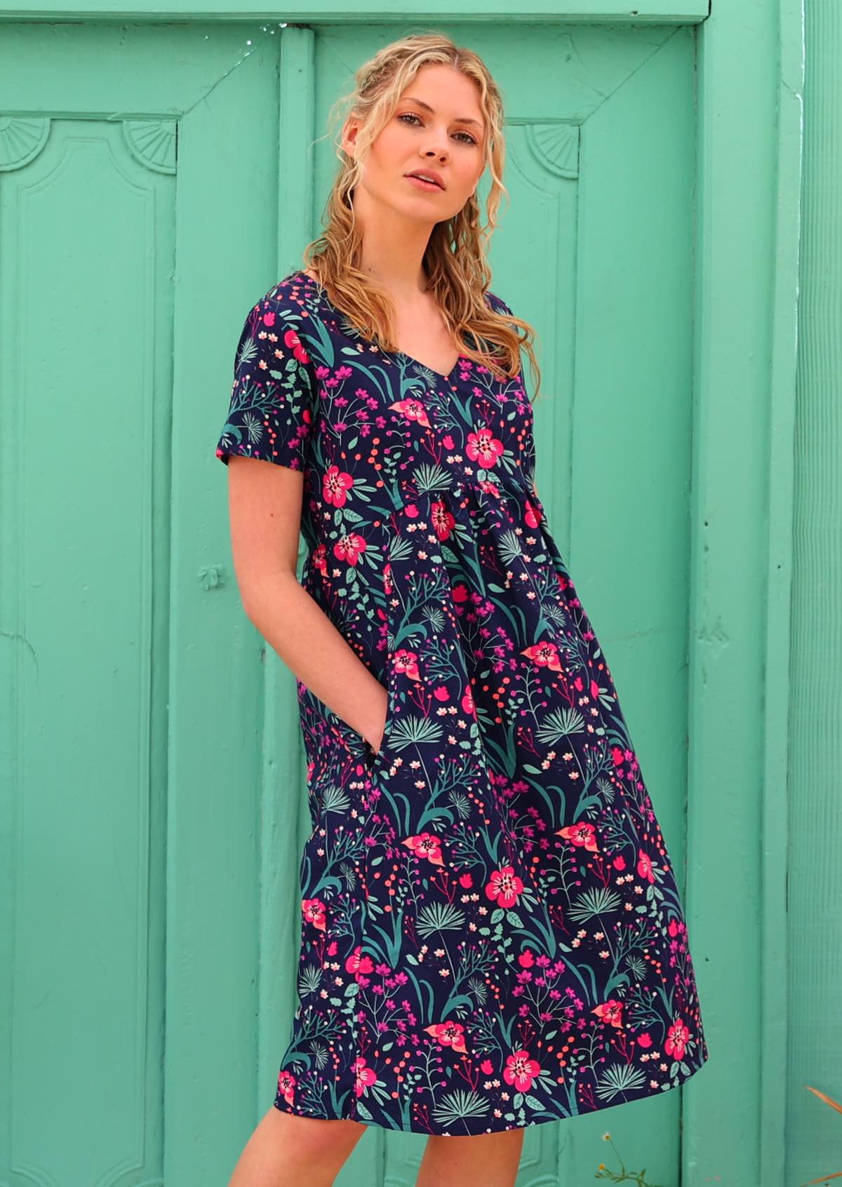 Model with hands in pockets wearing 100 percent cotton v-neck empire waist sundress with hot pink and bright purple flowers on a deep blue base 