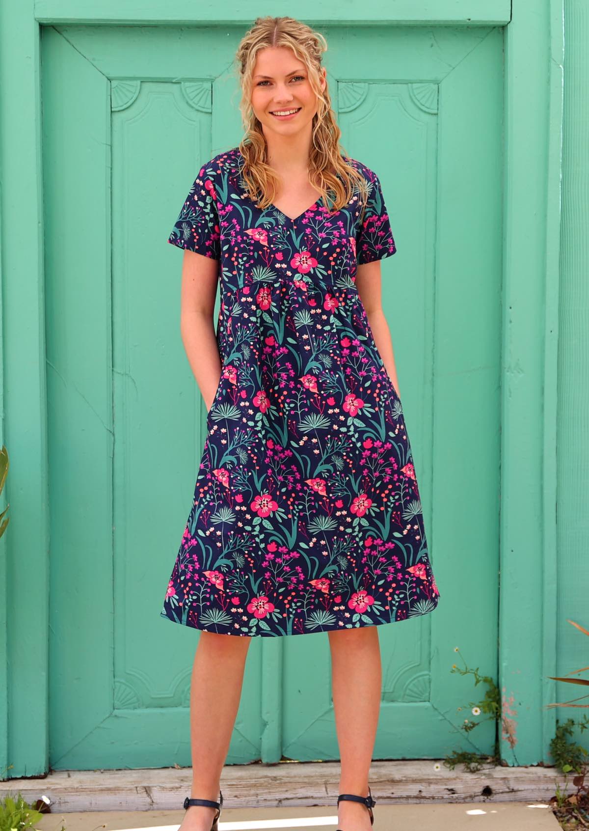 Model wearing 100 percent cotton v-neck sundress with hot pink and bright purple flowers on a deep blue base, with hands in pockets 