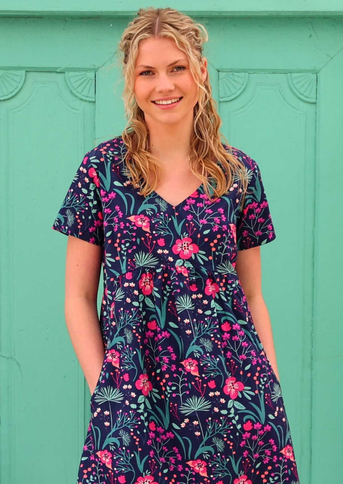 Close up of model with hands in pockets wearing 100 percent cotton v-neck empire waist sundress with hot pink and bright purple flowers on a deep blue base 