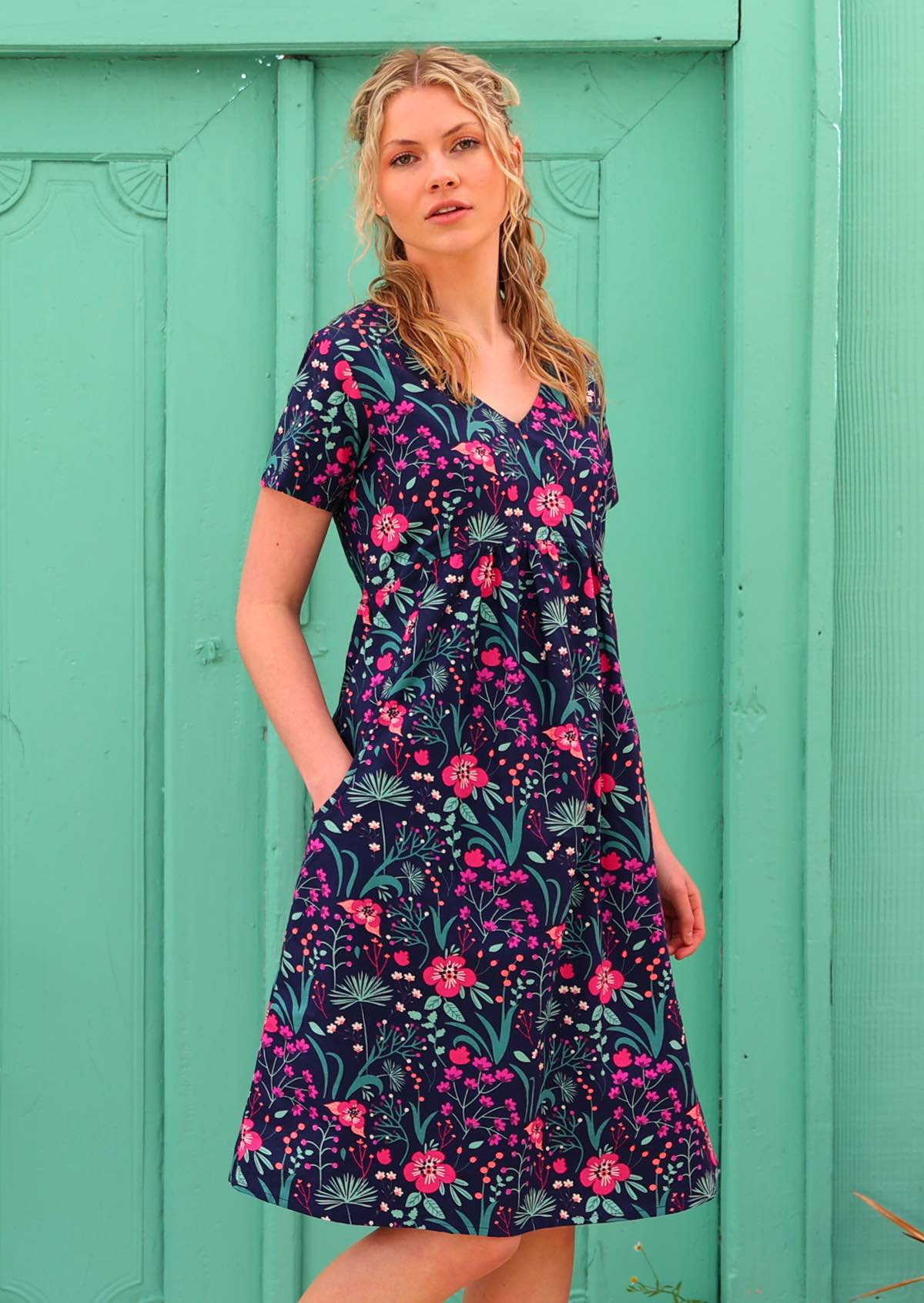 Model with one hand in pockets wearing 100 percent cotton v-neck empire waist sundress with hot pink and bright purple flowers on a deep blue base 