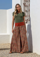 Woman wearing a green top and wide legged patterned cotton pants standing in front of a white wall with a textured design.