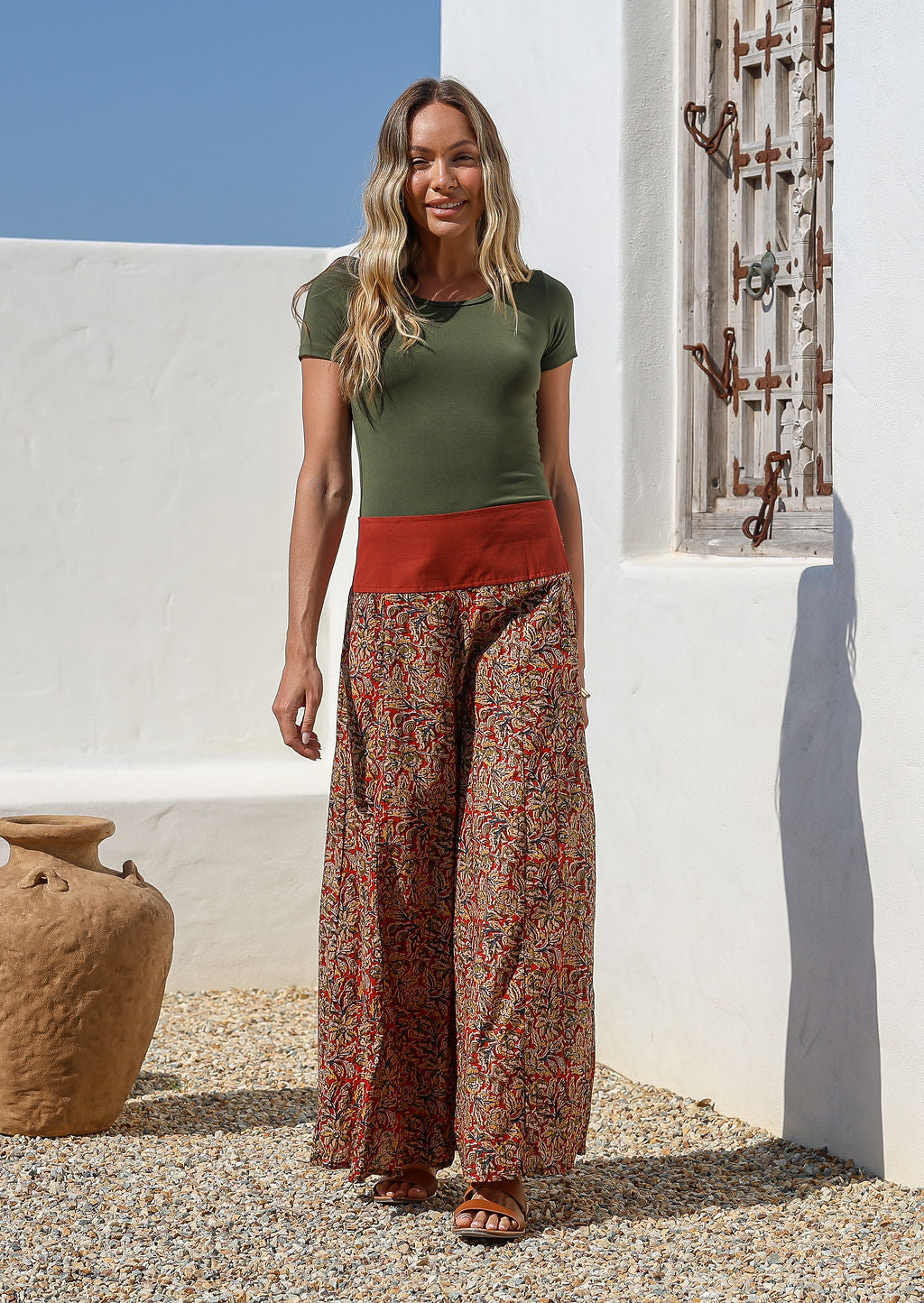 Woman wearing a green top and wide legged patterned cotton pants standing in front of a white wall 
