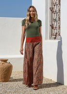 Woman wearing a green top and wide legged patterned cotton pants standing in front of a white wall 
