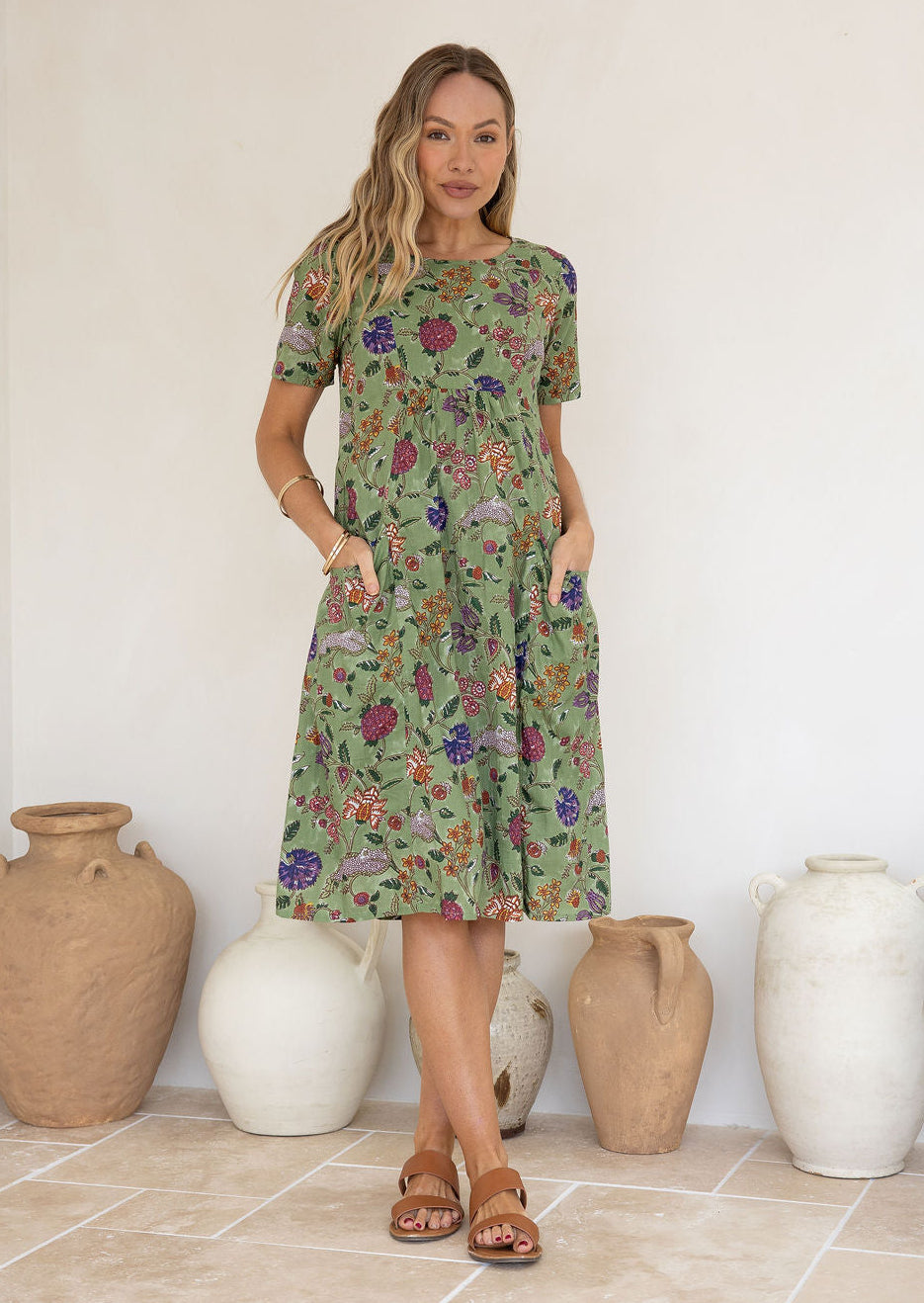 Woman wearing a green based floral print cotton dress with hands in pockets.