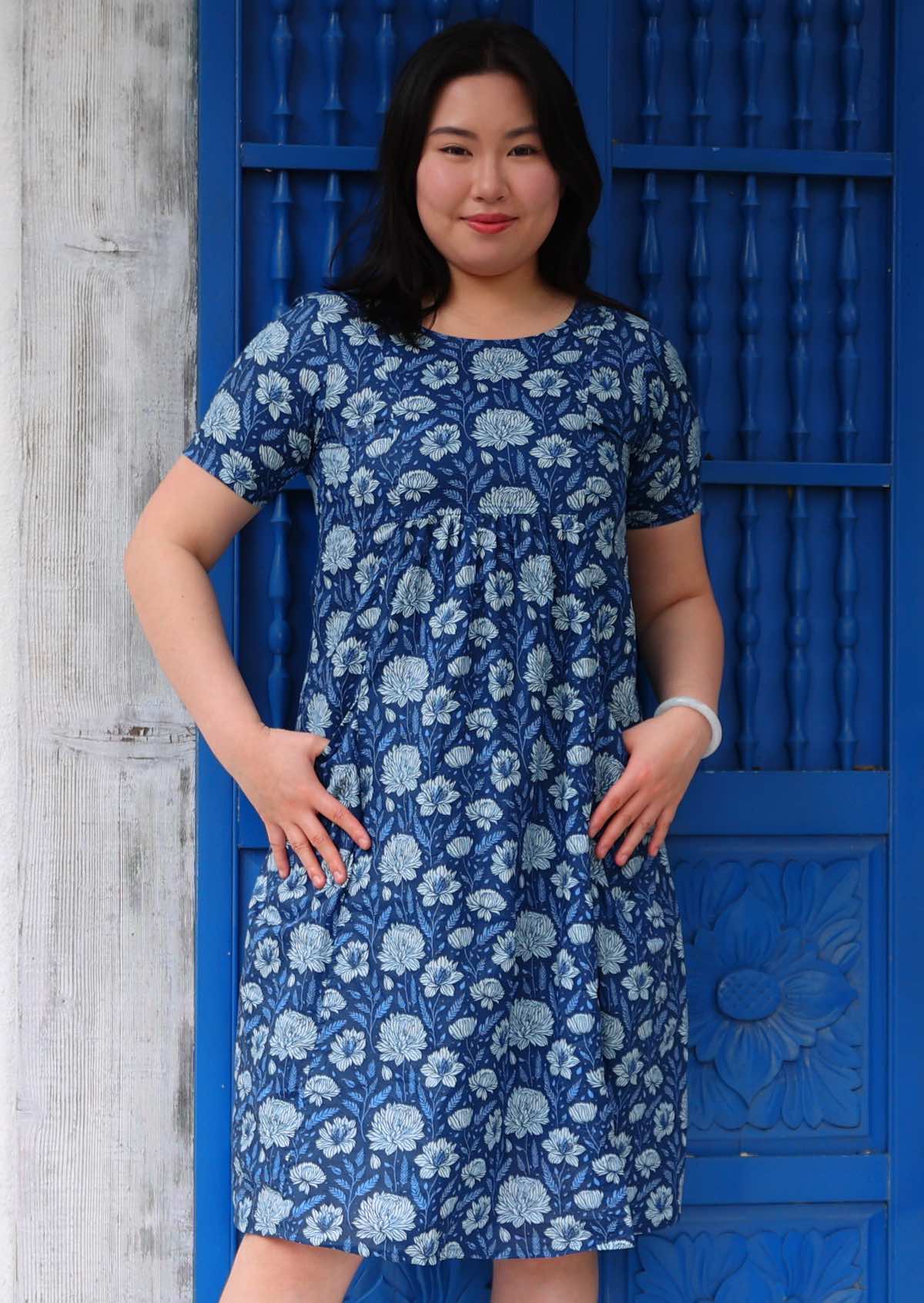 Woman with dark hair wearing a blue cotton dress with short sleeves and round neckline in light blue and white floral design, in front of blue door