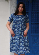 Woman with dark hair wearing a blue cotton dress with light blue and white floral design, in front of blue door, with her thumbs in pockets