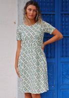 Woman wearing white cotton dress with floral pattern in front of bright blue door with hand on ྀིྀི