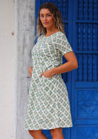 Woman wearing white cotton dress with floral pattern in front of bright blue door with hands in pockets 