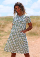 Woman wearing white cotton sundress with floral pattern on grassy hill with hands in pockets 