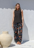 Woman standing in front of white wall wears 100% cotton wide leg botanic print pants in black with elastic waist, pockets and pleated ankles