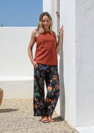 Woman wearing a rust-colored sleeveless top and dark botanic print wide leg cotton pants with pleated ankles, elastic waist and pockets