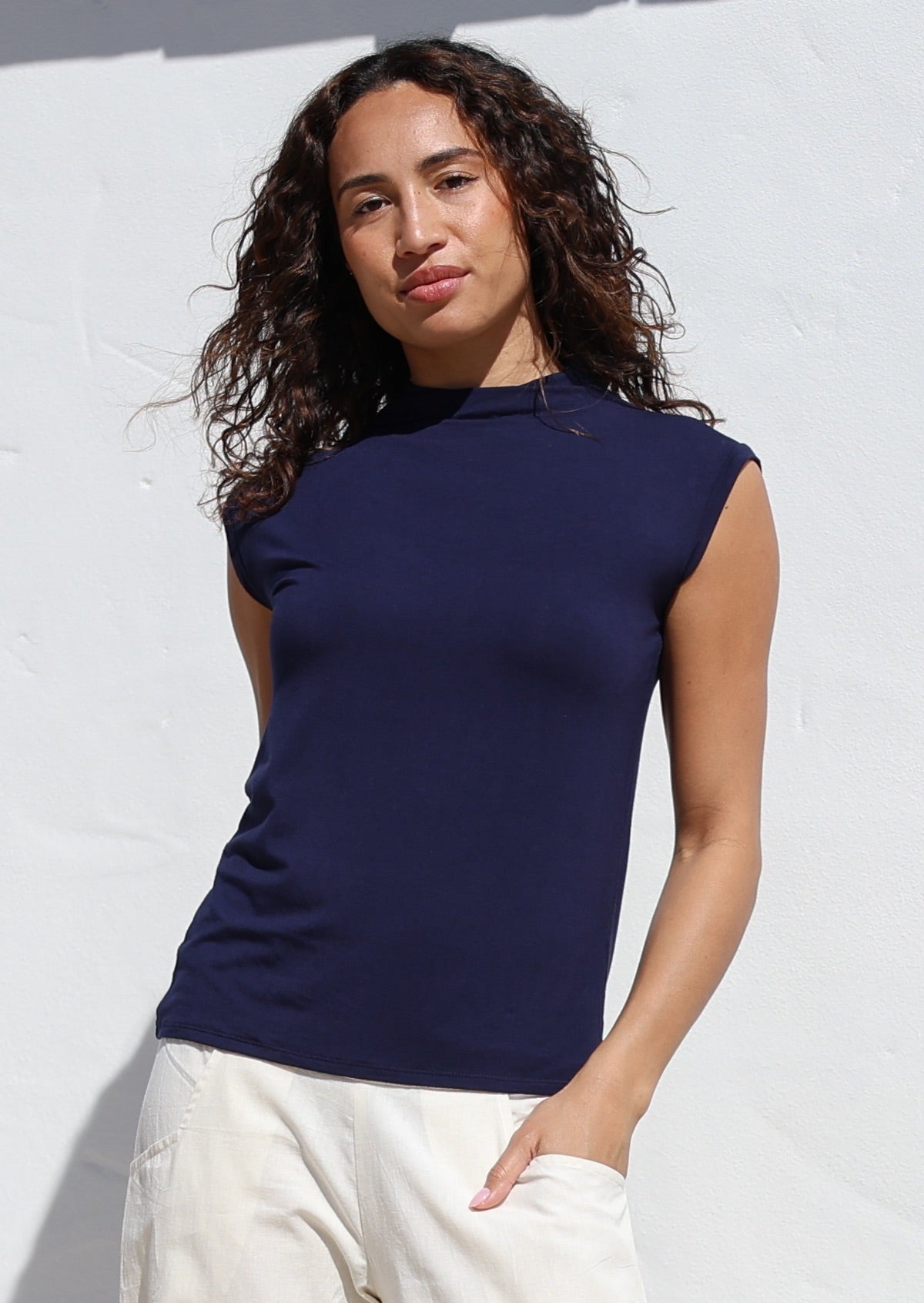 Woman wearing a navy sleeveless top against a white background, with hand in pants pocket