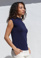 Woman wearing a navy sleeveless top with a high neckline, with hands in pockets of white pants