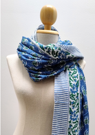 A blue patterned scarf with green floral motifs and white stripes, draped on a cream-colored fabric mannequin torso with a wooden neck cap.