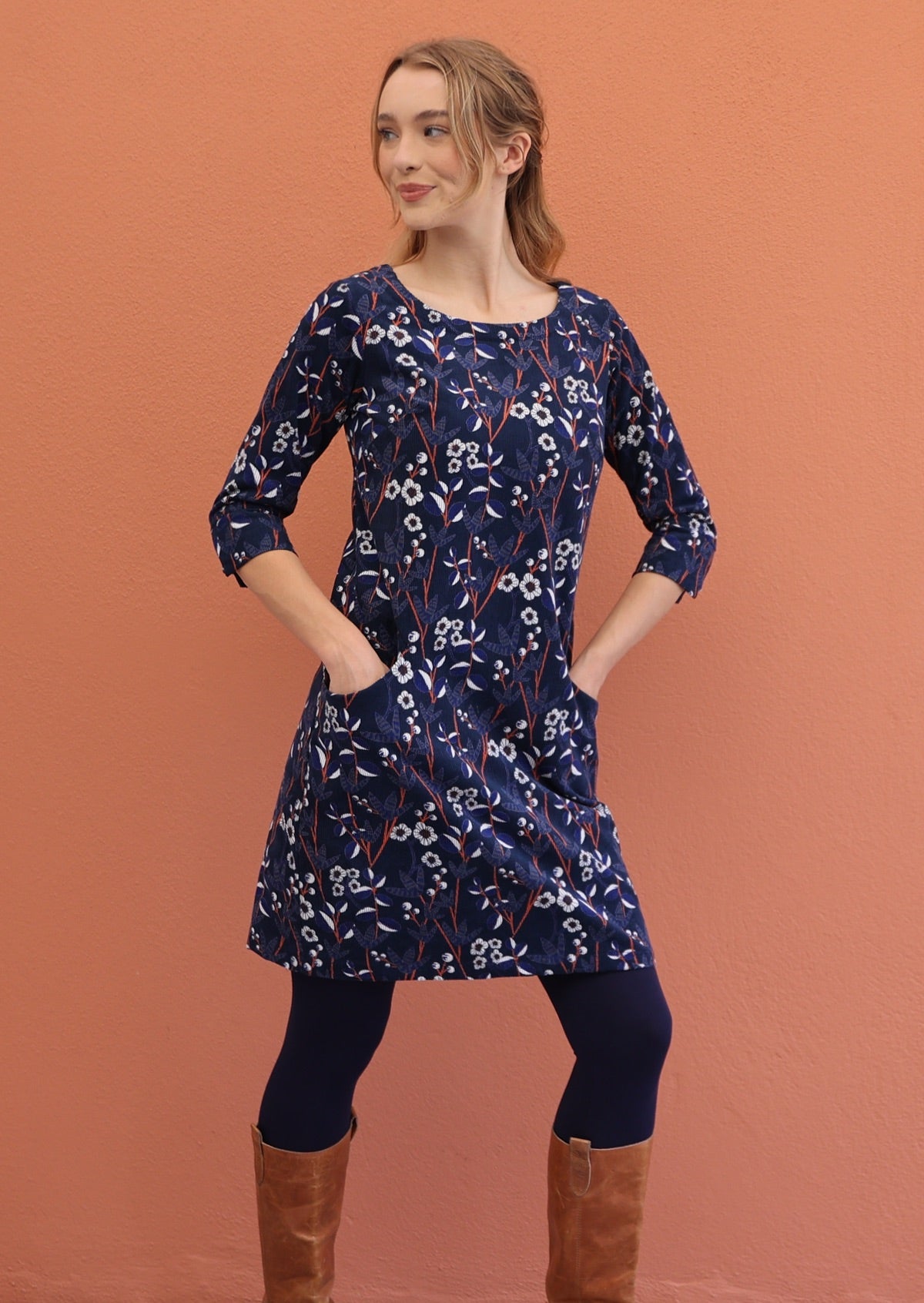 Woman wears  round neck, 3/4 sleeve corduroy dress with hands in pockets, paired with navy blue leggings and boots