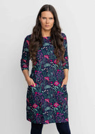 Woman leaning against wall wearing Cotton 3/4 sleeve shift dress with detailed cuff, in gorgeous floral print on dark blue base over navy blue leggings 