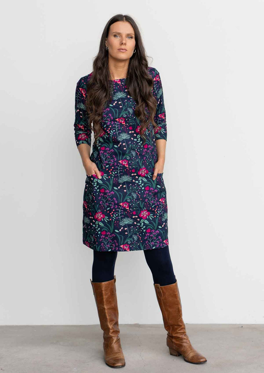Woman leaning against wall wearing Cotton 3/4 sleeve shift dress with detailed cuff, in gorgeous floral print on dark blue base over navy blue leggings and tan boots 