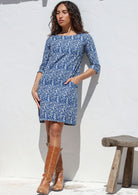 Woman wearing a blue cotton floral dress with brown boots standing against a white wall.