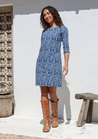 Woman wearing a blue cotton dress with brown boots standing against a white wall.
