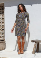 Woman wearing a black cotton patterned dress and brown boots standing against a white wall.