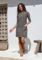 Woman wearing a cotton black patterned dress and brown boots standing against a white wall.