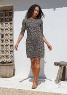 Woman wearing a black floral cotton patterned dress and brown boots standing against a white wall.