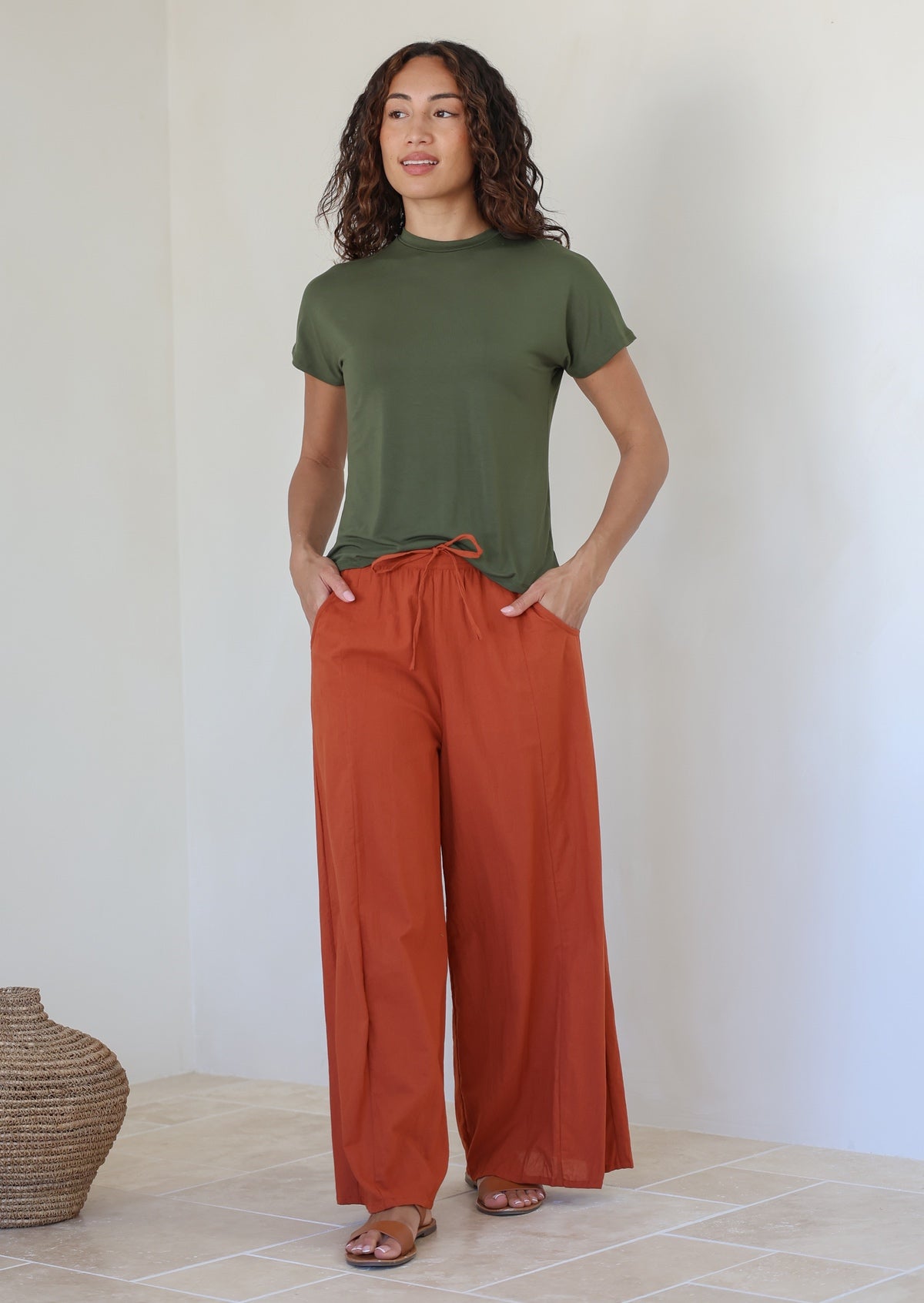 Woman wearing lightweight 100% cotton wide leg pants in brick orange shade with pockets, drawstring tie and elastic at back of the waist