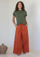 Woman wearing a green t-shirt and rust colored wide-leg cotton pants with drawstring tie and hands in pocket standing against the white wall.