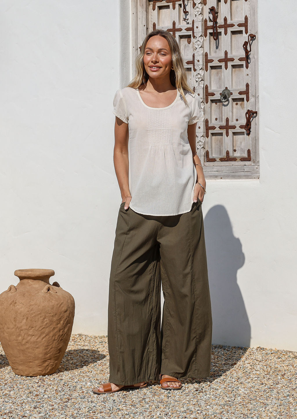 Woman wearing a white cotton top and olive green wide leg cotton pants standing in front of a white wall with a wooden window