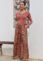 Woman stands in cotton wide leg pants in earthy toned floral print on red base.