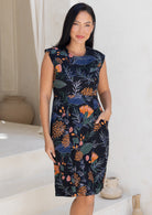Woman wearing a sleeveless dress with a black based floral print, with one hand in front pocket
