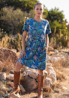 Woman with one foot on a rock wearing 100 percent cotton blue base retro dress with leafy print with one hand in pocket