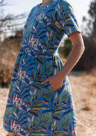 close up of woman wearing 100 percent cotton blue base retro dress with leafy print and short sleeves with hand in pocket