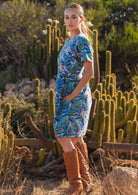 Woman standing to the side facing forward wearing 100 percent cotton blue base retro dress with leafy print with one hand in pocket