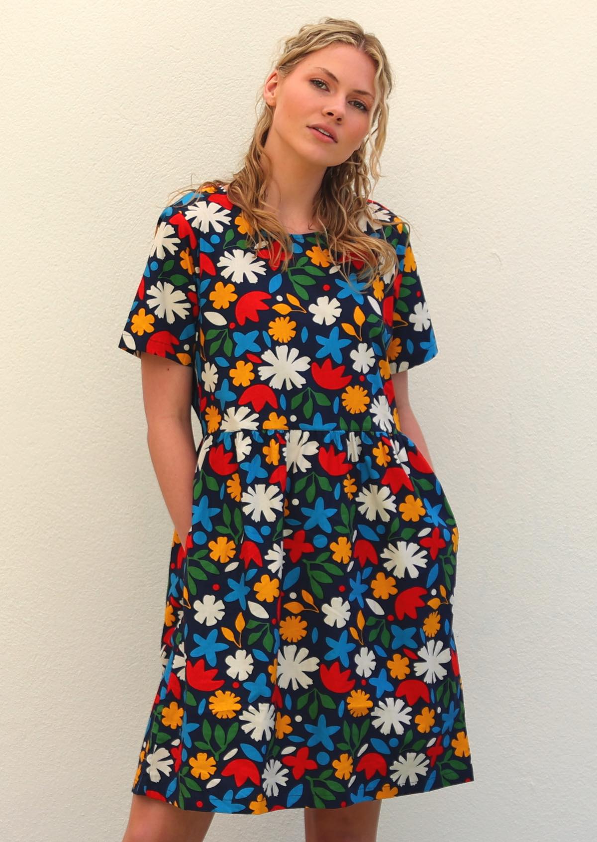 Model with blonde hair wearing 100 percent cotton dark navy blue base and bright abstract floral retro print cotton dress, with hands in deep side pockets 