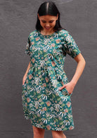 Model with dark hair in loose fit green cotton floral dress in front of dark grey wall