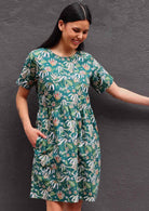 Model with dark hair in loose fit green cotton floral dress with  pockets in front of dark grey wall