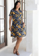 Woman stands with hand in pocket wearing a 100% cotton relaxed fit dress with round neckline and short sleeves in black based retro floral print