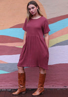 Full length photo pf model wearing 100% cotton short sleeve dress in plum colour with hands in deep pockets