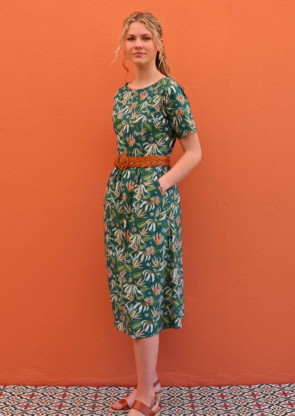 Model standing side on with hands in pockets wearing 100 percent green cotton sundress with sleeves and waist belt