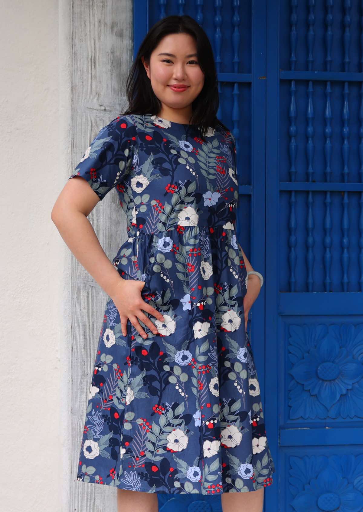 Woman stands on the side with thumbs in side pockets of cotton midi length dress in blue floral print with pops of red